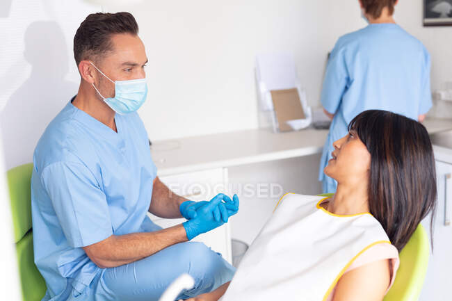 Dentista masculino caucásico con máscara facial hablando con una paciente femenina en una clínica dental moderna. negocio de salud y odontología. - foto de stock