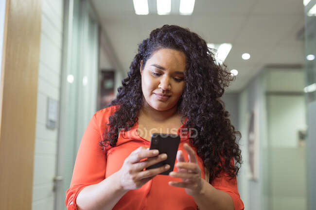 Femme d'affaires biraciale souriante marchant et utilisant un smartphone dans le couloir dans un bureau moderne. lieu de travail professionnel et de bureau. — Photo de stock