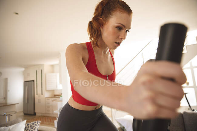 Mulher caucasiana passar o tempo em casa, na sala de estar, exercitando-se em cross trainer, vestindo roupas esportivas. — Fotografia de Stock