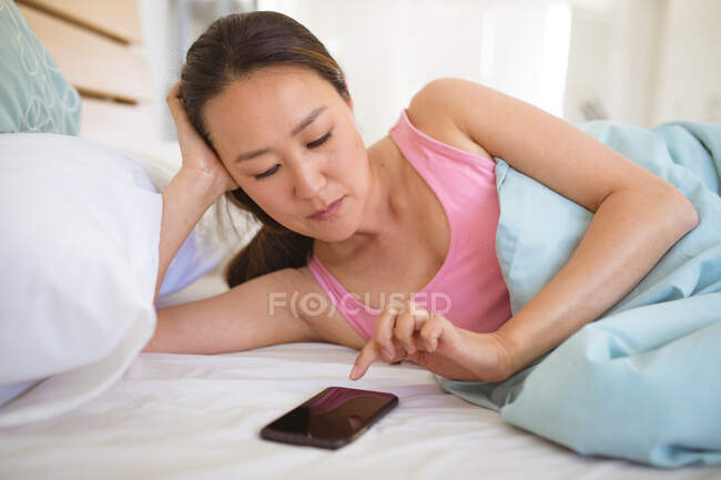 Focada mulher asiática deitada na cama, descansando e usando smartphone. relaxante em casa com a tecnologia. — Fotografia de Stock