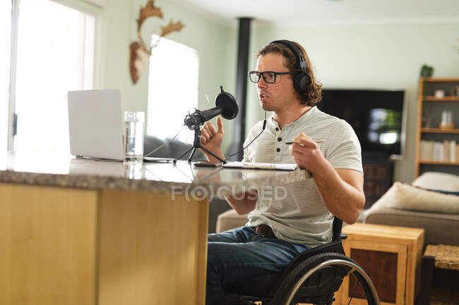 Kaukasischer behinderter Mann, der einen Podcast mit einem Mikrofon aufzeichnet, der zu Hause im Rollstuhl sitzt. Blogging, Podcast und Sendetechnologie-Konzept — Stockfoto