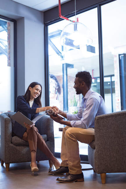 Empresario afroamericano sosteniendo portapapeles y hablando con una colega en una oficina moderna. - foto de stock