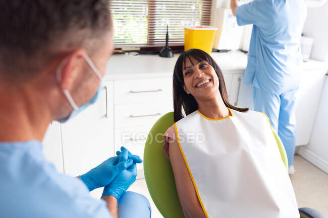 Dentista masculino caucásico con máscara facial hablando con una paciente femenina sonriente en una clínica dental moderna. negocio de salud y odontología. - foto de stock