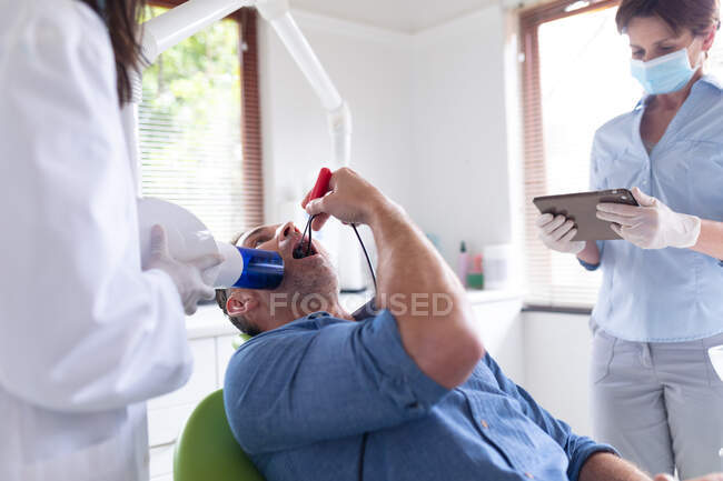 Dentista feminina com enfermeira dentária caucasiana examinando dentes de paciente do sexo masculino na clínica odontológica moderna. serviços de saúde e odontologia. — Fotografia de Stock