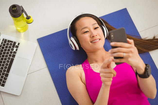 Felice donna asiatica con le cuffie sdraiata sul tappeto, che si esercita a casa con lo smartphone. sano stile di vita attivo e fitness a casa con la tecnologia. — Foto stock