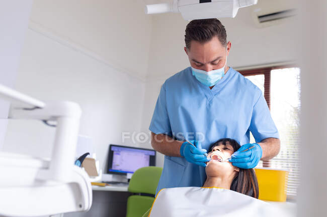 Dentista masculino caucásico que usa mascarilla facial examinando los dientes de una paciente femenina en una clínica dental moderna. negocio de salud y odontología. - foto de stock