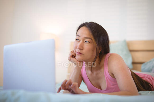 Nachdenkliche Asiatin, die auf dem Bett liegt, sich ausruht und das Smartphone benutzt. Mit Technik zu Hause entspannen. — Stockfoto