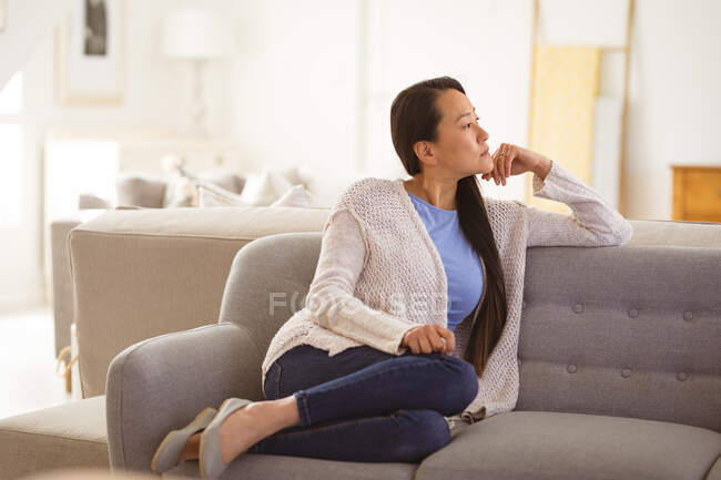 Задумчивая азиатка, сидящая дома на диване. образ жизни, отдых и проведение времени дома. — стоковое фото
