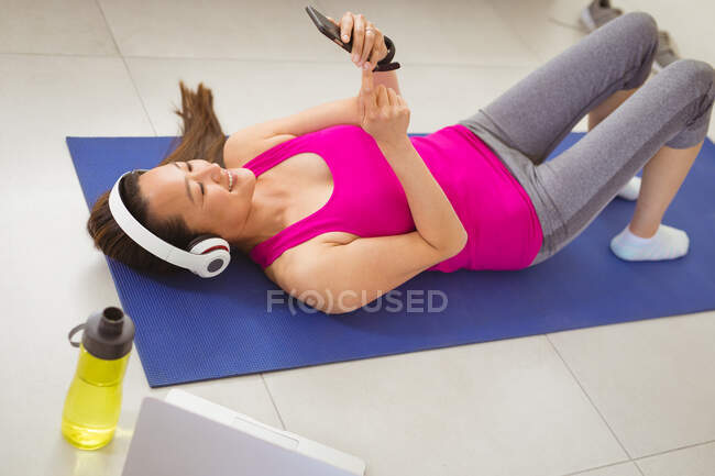 Mujer asiática feliz con auriculares acostados en la estera, haciendo ejercicio en casa con teléfono inteligente. estilo de vida activo saludable y fitness en el hogar con tecnología. - foto de stock