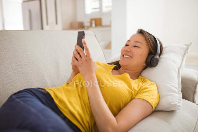 Heureuse femme asiatique portant des écouteurs allongés sur un canapé avec smartphone à la maison. style de vie et détente à la maison avec la technologie. — Photo de stock
