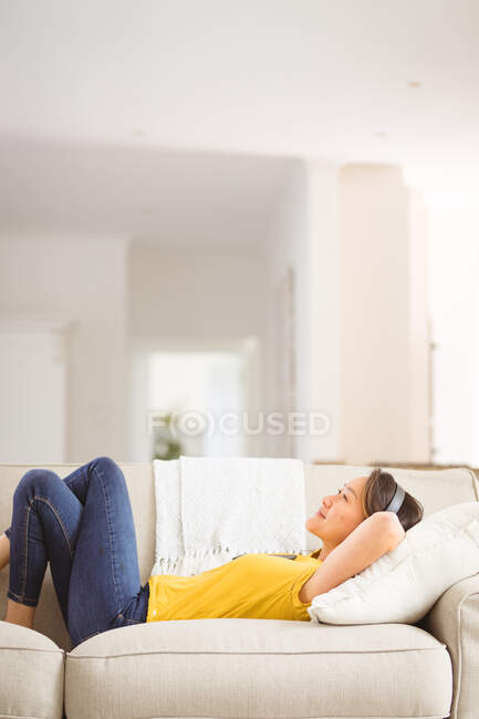 Feliz mulher asiática usando fones de ouvido descansando no sofá em casa. estilo de vida e relaxar em casa com a tecnologia. — Fotografia de Stock