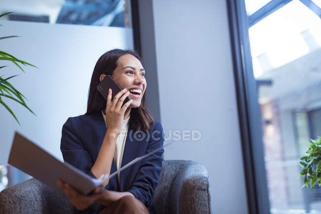 Mujer de negocios Biracial sonriendo, sosteniendo documentos y hablando en el teléfono inteligente en la oficina moderna. negocios y oficina de trabajo. - foto de stock