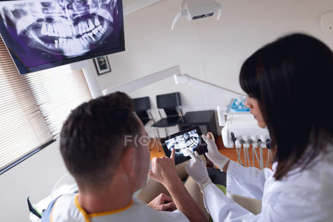 Odontóloga Biracial usando máscara facial examinando los dientes de un paciente masculino en una clínica dental moderna. negocio de salud y odontología. - foto de stock