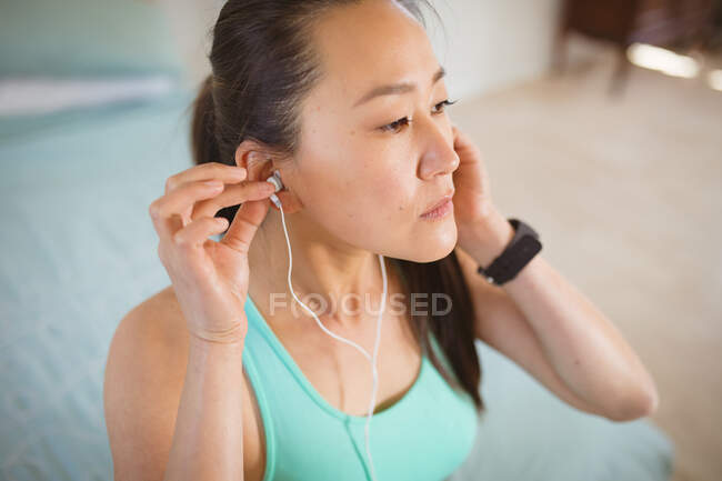 Retrato de mujer asiática en ropa de fitness, preparándose para el ejercicio, poniéndose auriculares. estilo de vida activo saludable y aptitud física en casa. - foto de stock
