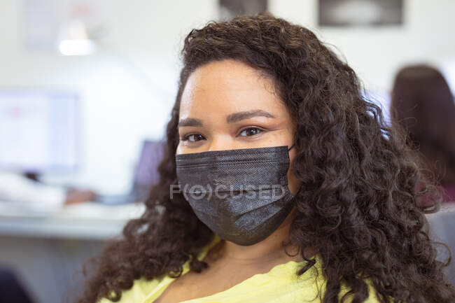 Retrato de una mujer de negocios birracial sonriente con máscara facial mirando a la cámara en la oficina moderna. negocio y oficina durante la pandemia de covid 19. - foto de stock