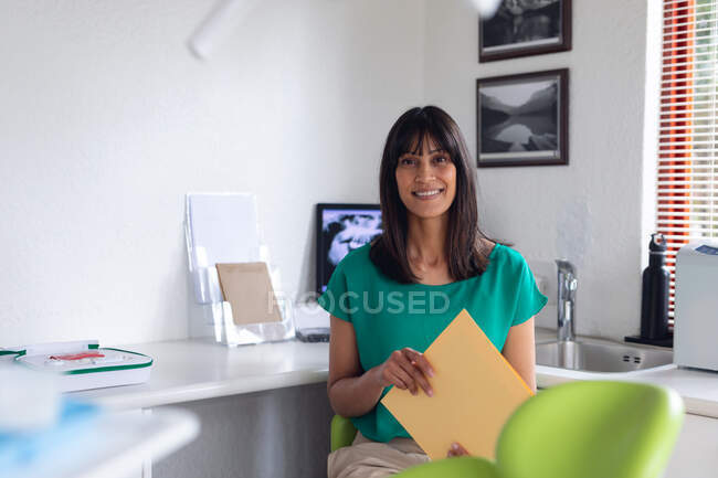 Retrato de una paciente birracial sonriente sosteniendo documentos en una clínica dental moderna. negocio de salud y odontología. - foto de stock