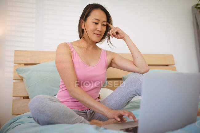 Задумчивая азиатка, сидящая на кровати, отдыхающая и пользующаяся смартфоном. расслабляющий дома с технологией. — стоковое фото