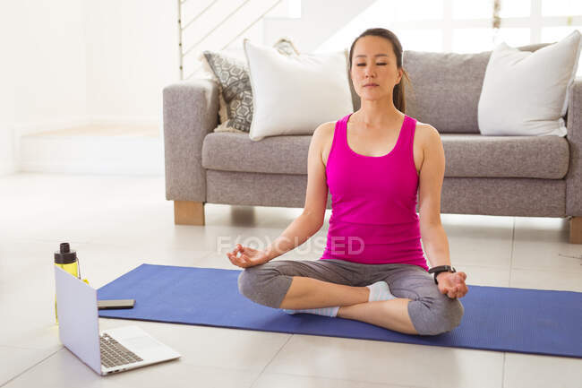 Зосереджена азіатська жінка сидить на килимку, роздумуючи вдома з ноутбуком. здоровий активний спосіб життя і фізична підготовка вдома з технологіями . — стокове фото