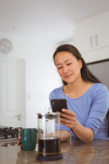 Heureuse femme asiatique buvant du café et utilisant smartphone dans la cuisine. style de vie et détente à la maison avec la technologie. — Photo de stock