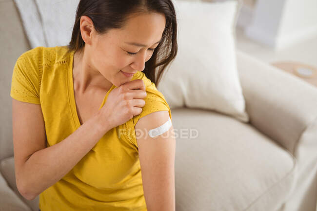 Felice donna asiatica seduta sul divano mostrando braccio con gesso dopo la vaccinazione. salute e stile di vita durante la pandemia della congrega 19. — Foto stock