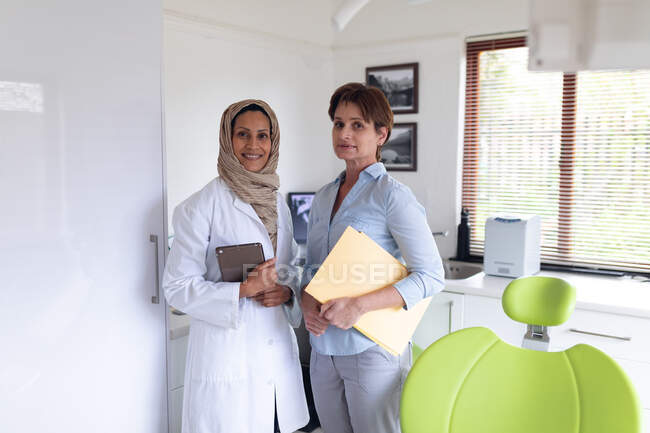 Retrato de sorriso biracial feminino dentista e enfermeira dentária feminina na clínica odontológica moderna. serviços de saúde e odontologia. — Fotografia de Stock