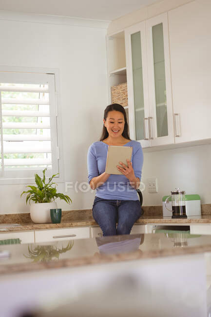 Heureuse femme asiatique assise sur le contour et en utilisant une tablette dans la cuisine. style de vie et détente à la maison avec la technologie. — Photo de stock