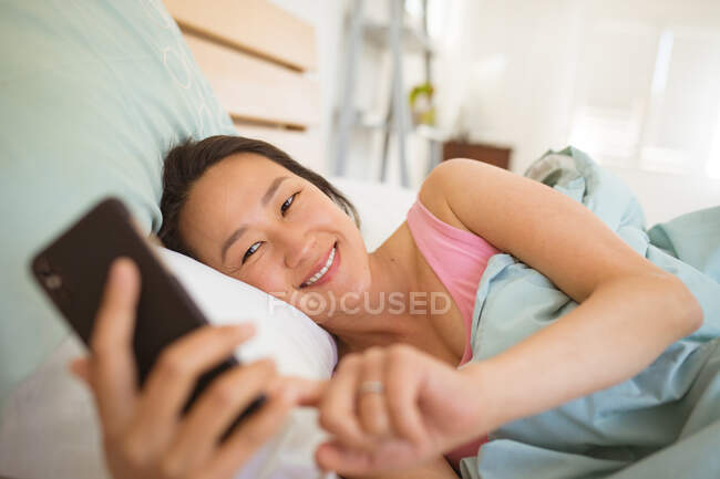 Щаслива азіатка лежить на ліжку, відпочиває і використовує смартфон. розслабляючись вдома з технологією . — стокове фото