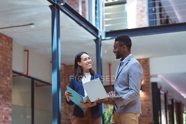 Two diverse business people smiling and talking in modern office with colleagues in background. business and office workplace. — Stock Photo