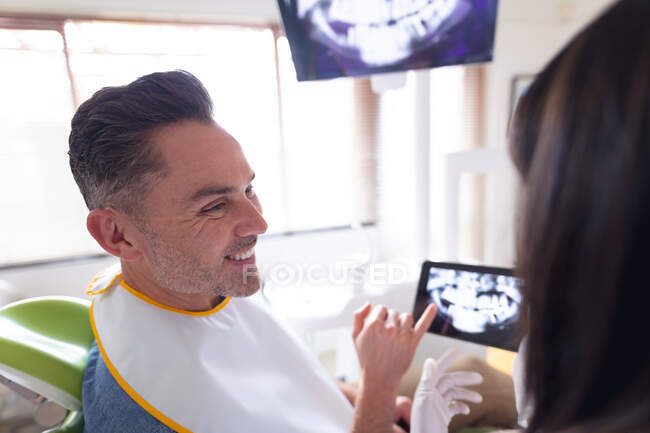 Dentiste féminine portant des gants et examinant les dents d'un patient souriant de sexe masculin à la clinique dentaire moderne. — Photo de stock