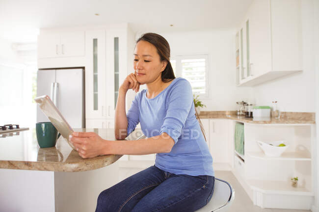 Счастливая азиатка сидит за столом, пьет кофе и пользуется планшетом на кухне. образ жизни, отдых и проведение времени дома с технологиями. — стоковое фото