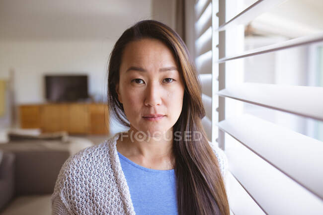 Porträt einer ernsthaften asiatischen Frau, die am Fenster steht und in die Kamera blickt. Lebensstil, Freizeit und Zeit zu Hause verbringen. — Stockfoto