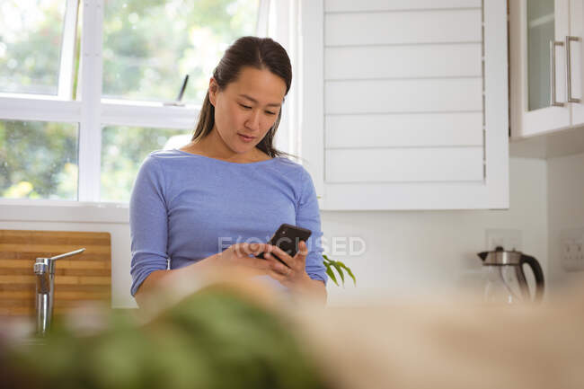 Mujer asiática enfocada usando smartphone en la cocina. estilo de vida, ocio y relax en casa con tecnología. - foto de stock