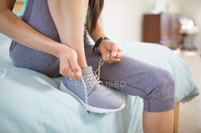 Мидсекция женщины, сидящей на кровати в спортивной одежде, готовящейся к упражнениям, завязывающей обувь. здоровый активный образ жизни и фитнес дома. — стоковое фото