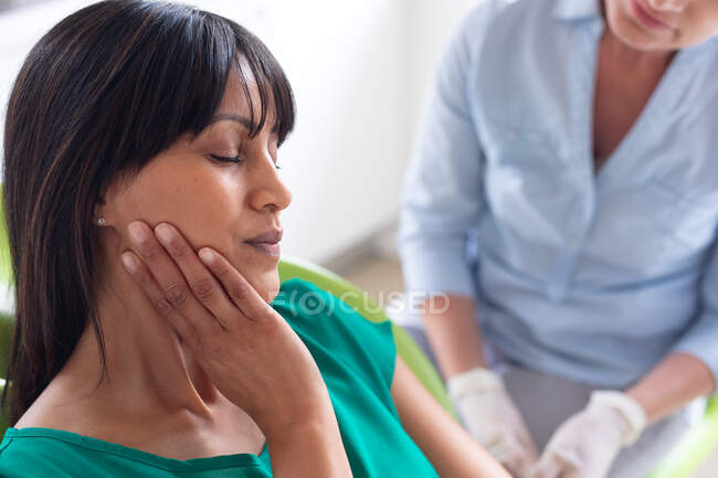 Biracial female patient holding her face with caucasian female dental nurse at modern dental clinic. healthcare and dentistry business. — Stock Photo