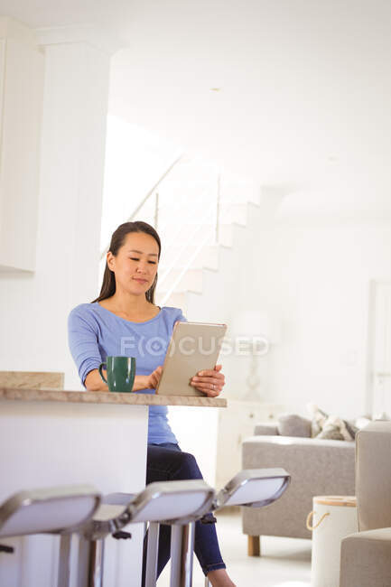 Asian woman sitting at table, drinking coffee and using tablet in kitchen. lifestyle and relaxing at home with technology. — Stock Photo