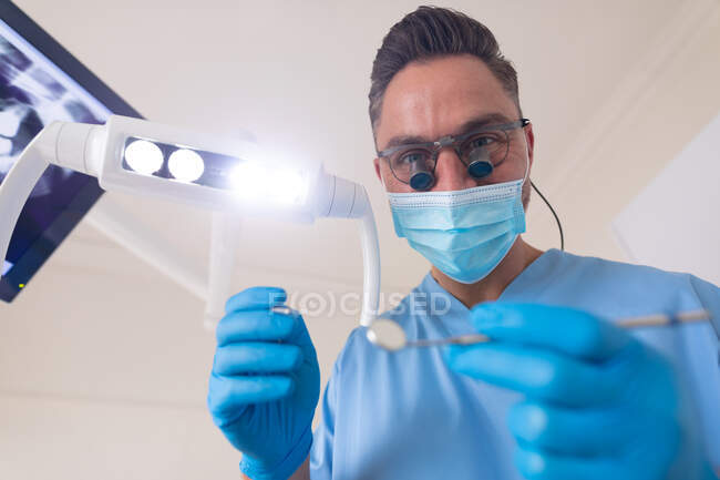 Homem dentista caucasiano usando máscara facial segurando ferramentas dentárias na clínica odontológica moderna. serviços de saúde e odontologia. — Fotografia de Stock