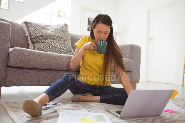 Focused asian woman drinking coffee and working remotely from home with smartphone and laptop. home office and freelancing concept. — Stock Photo