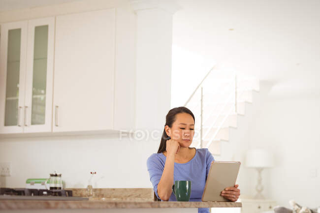 Азиатка, сидящая за столом, пьющая кофе и пользующаяся планшетом на кухне. стиль жизни и отдых дома с технологией. — стоковое фото