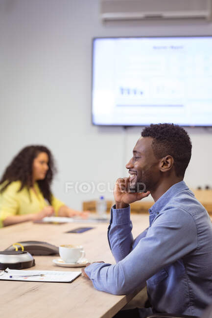 Homme d'affaires afro-américain souriant parlant sur smartphone et assis à table dans un bureau moderne. lieu de travail professionnel et de bureau. — Photo de stock