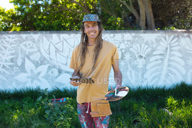 Retrato de artista masculino sorridente segurando paleta enquanto estava de pé pintura mural abstrata na parede. arte de rua e habilidade. — Fotografia de Stock