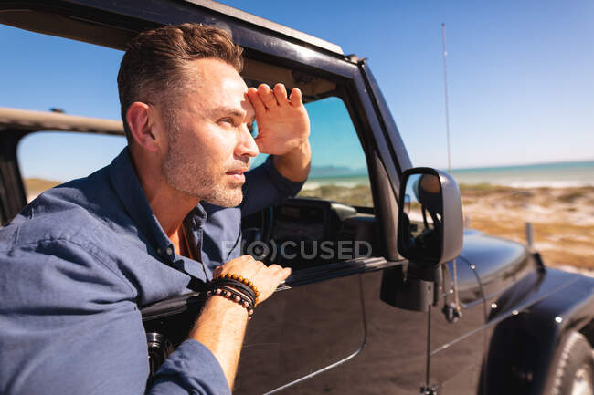 Thoughtful caucasian man sitting in car at seaside shielding eyes from sun and admiring the view. summer road trip and holiday in nature. — Stock Photo