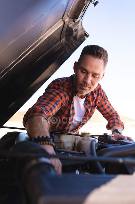 Focused caucasian man repairing car with bonnet open at seaside. summer road trip and holiday in nature. — Stock Photo