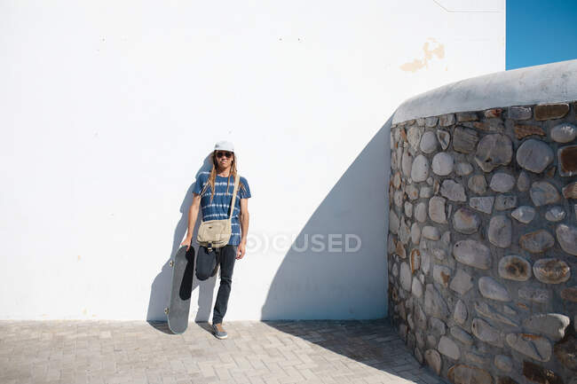 Longitud completa de hipster masculino sosteniendo monopatín mientras se apoya en la pared blanca en el día soleado. estilo de vida y deporte. - foto de stock