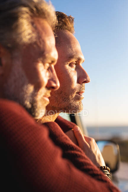 Feliz pareja de hombres caucásicos del mismo sexo abrazándose por un coche en el sol, disfrutando de la vista al mar. viaje por carretera de verano y vacaciones en la naturaleza. - foto de stock