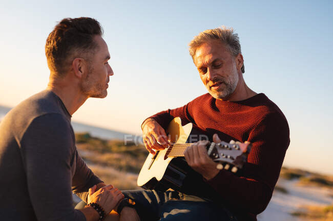 Felice caucasico gay coppia maschile appendere fuori sulla spiaggia al tramonto, suonare la chitarra. estate viaggio su strada e vacanza nella natura. — Foto stock
