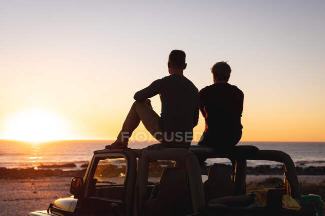 Rückansicht eines kaukasischen schwulen männlichen Paares, das bei Sonnenuntergang am Meer auf dem Autodach sitzt. Sommer Roadtrip und Urlaub in der Natur. — Stockfoto