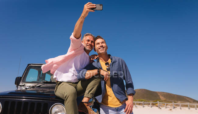 Feliz pareja gay caucásica abrazando y tomando selfies en un auto en la playa. viaje por carretera de verano y vacaciones en la naturaleza. - foto de stock