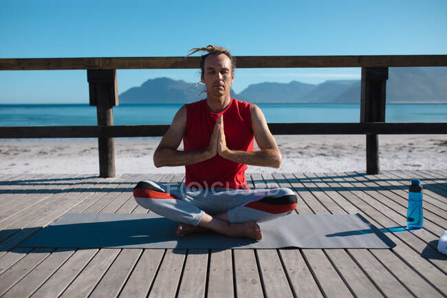 Man practicing yoga while sitting cross-legged and hands clasped meditating on floorboard at beach. fitness and healthy lifestyle. — Stock Photo