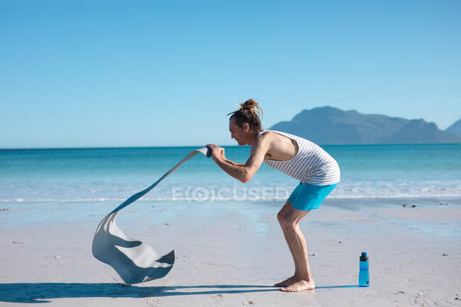 Vista lateral del hombre colocando esterilla de yoga para hacer ejercicio en la orilla de la playa durante el día soleado con espacio para copiar. fitness y estilo de vida saludable. - foto de stock