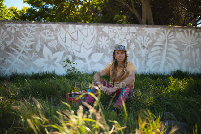 Retrato de artista masculino sentado na grama contra pintura mural abstrata na parede. arte de rua e habilidade. — Fotografia de Stock